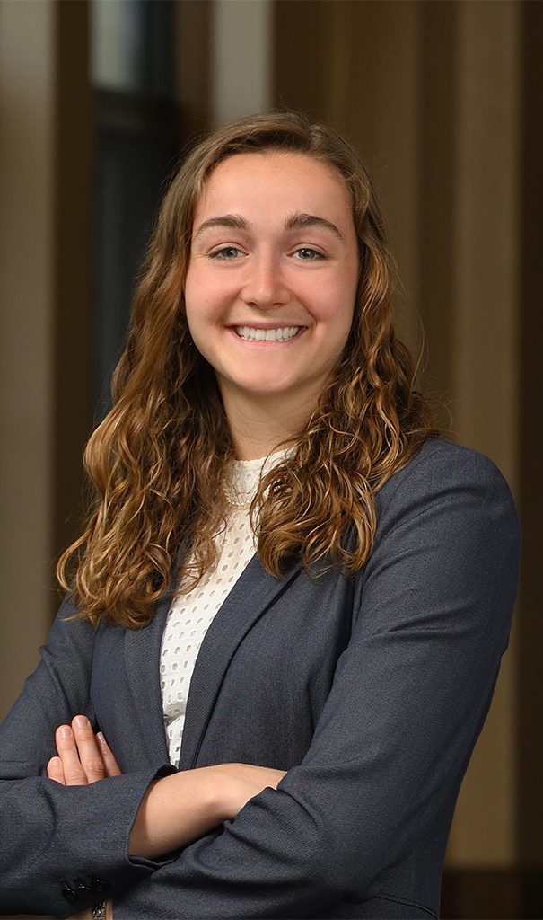 woman with curly brown hair smiling at the camera, she is wearing a blazer over a sweater