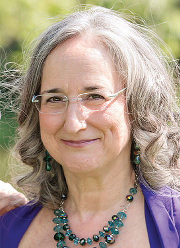 Photo of Ellen Pollak. Older woman with shoulder length curled grey hair, wearing glasses, a blue beaded necklace and a purple blouse.