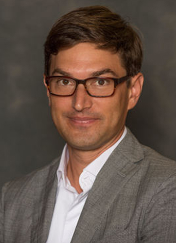 Photo of Kaveh Askari. Middle aged man with short brown hair, and glasses, and grey suit coat.