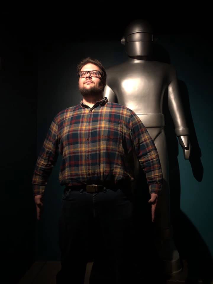 A man in a dark room wearing a plaid shirt standing in front of a robot statue and posing in a similarly statuesque manner.