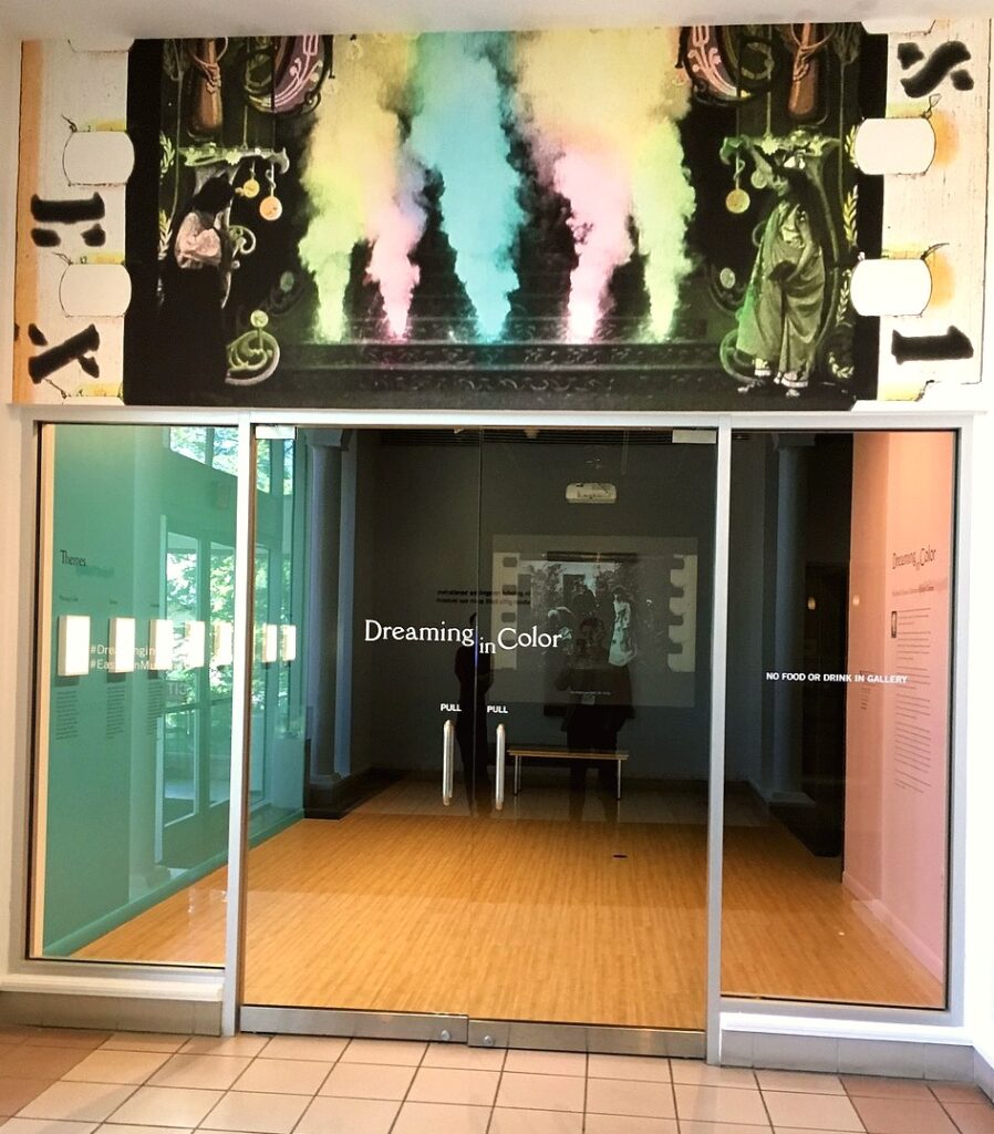 Glass entrance doors to an art gallery with the exhibit title "Dreaming in Color" displayed in white lettering. Above the doors is a vivid film still featuring black-and-white figures standing beside billowing clouds of pastel-colored smoke. Exhibition walls and panels are visible inside the gallery space.