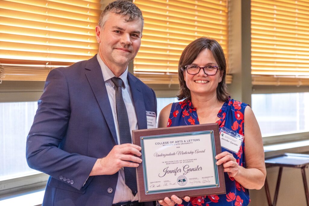Two people stand together in a room with windows and blinds. The person on the right, wearing glasses and a blue and red flowered top, smiles while holding a framed plaque. On the left stands a man in a navy suit and tie that holds the other side of the plaque. The award reads, “Undergraduate Mentorship Award – Jennifer Gansler, College of Arts & Letters, Michigan State University.”