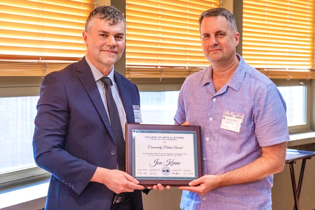 Two men stand indoors, smiling. The man on the left, in a navy suit and tie, holds one side of a framed award plaque, while the man on the right, in a short-sleeve lavender button-down shirt, holds the other side. The plaque reads, “Community Partner Award – Jon Keune, College of Arts & Letters, Michigan State University.”