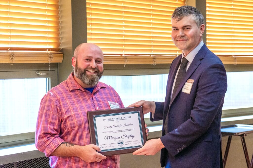 Two men stand side by side indoors. The man on the left, wearing a pink plaid shirt, smiles broadly while holding a framed award plaque. The man on the right, in a navy suit and tie, holds the other side of the plaque. The award reads, “Faculty Award for Innovation – Morgan Shipley, College of Arts & Letters, Michigan State University.”