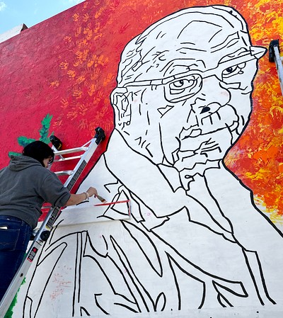 An artist stands on a ladder, painting details on the large mural of an older man outlined in black against a red and yellow background.