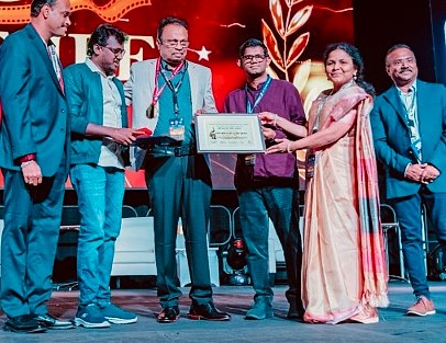A group of six people stand on stage during an award ceremony. A woman in a cream and red sari presents a framed certificate to a man at the center of the group, while others look on and smile. The background features a large, illuminated event display