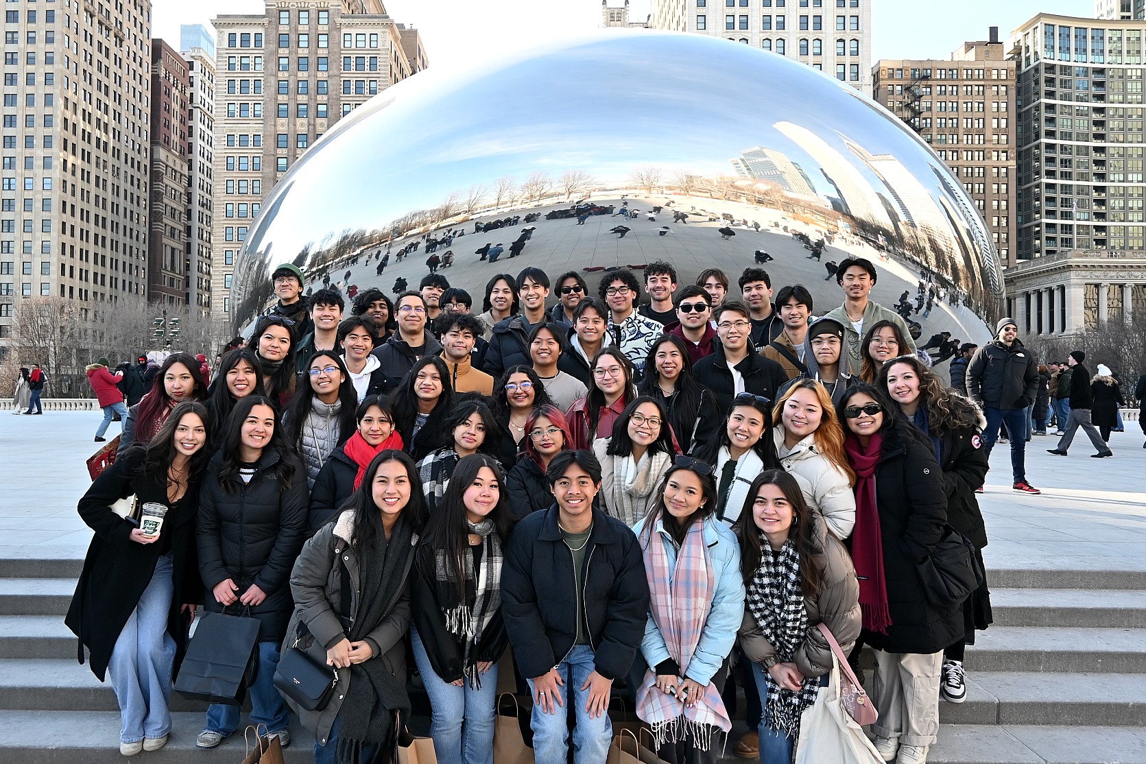 A large group of approximately forty students poses for a group photo in front of "Cloud Gate," also known as The Bean, in Chicago’s Millennium Park. The students are dressed in winter attire, including coats and scarves. The city’s skyline and the reflection of the plaza are visible on the sculpture's polished steel surface.