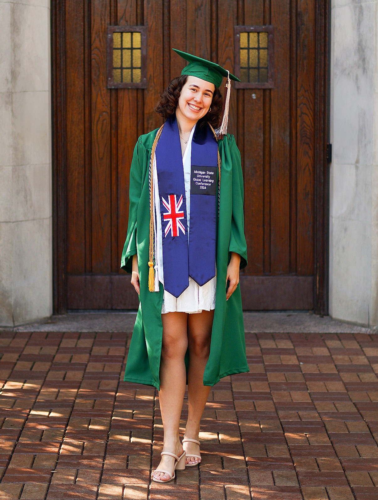 Young graduate in a green cap and gown stands smiling in front of wooden doors. She wears a blue sash with a Union Jack, exuding pride and joy.