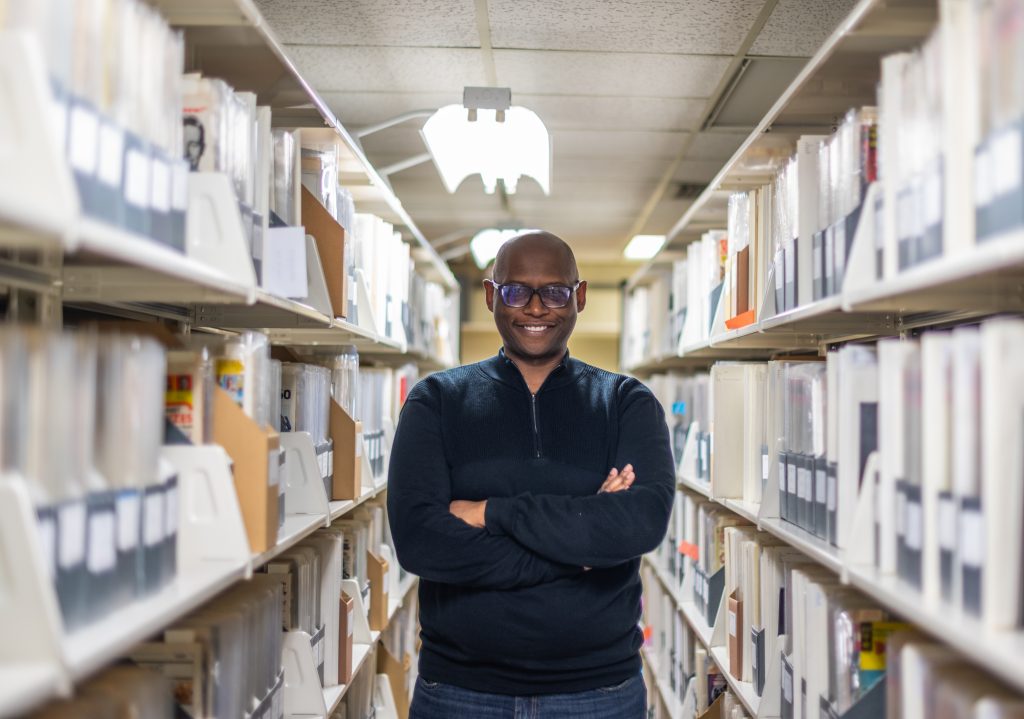 Man with crossed arms and blue sweater, standing in an aisle of books, looking at the camera smiling. 