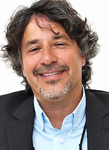 Photo of John J. Valadez. Middle aged man with curly black and grey hair. Wearing blue shirt and black suit coat.