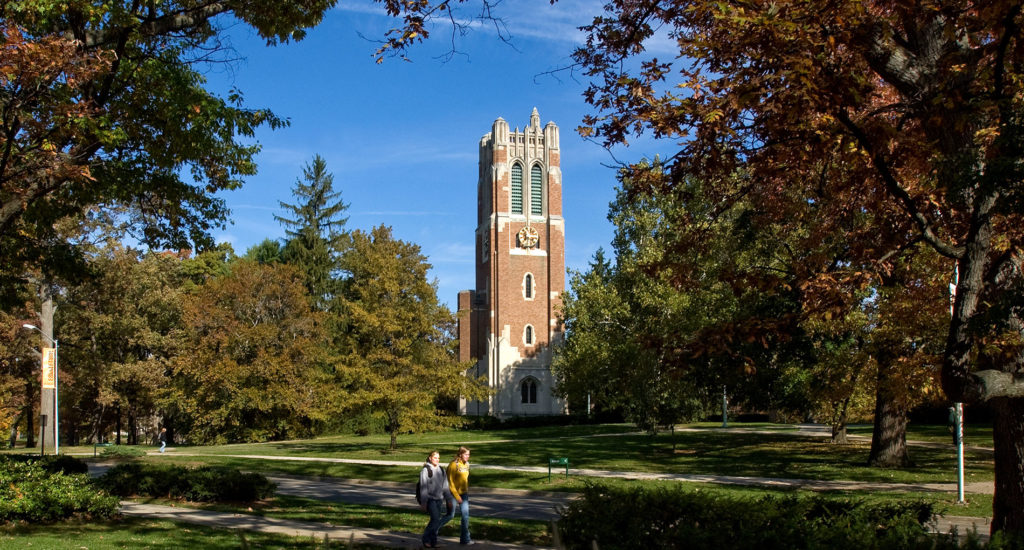 beaumont tower in the background surrounded by trees and greenery and two people walking in the distance