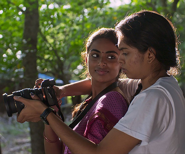 Two women holding a camera. The woman on the right wears a white t-shirt and has her arm around the other woman. The woman on the left is wearing a pink shirt with blue polka dots and is holding the camera.