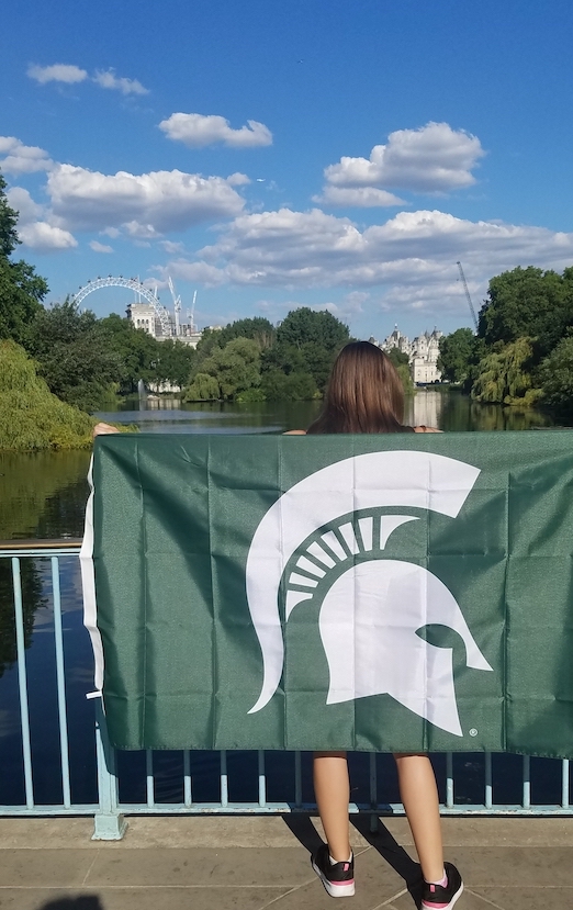 woman with long brown hair holding up a Michigan state flag facing water and blue sky