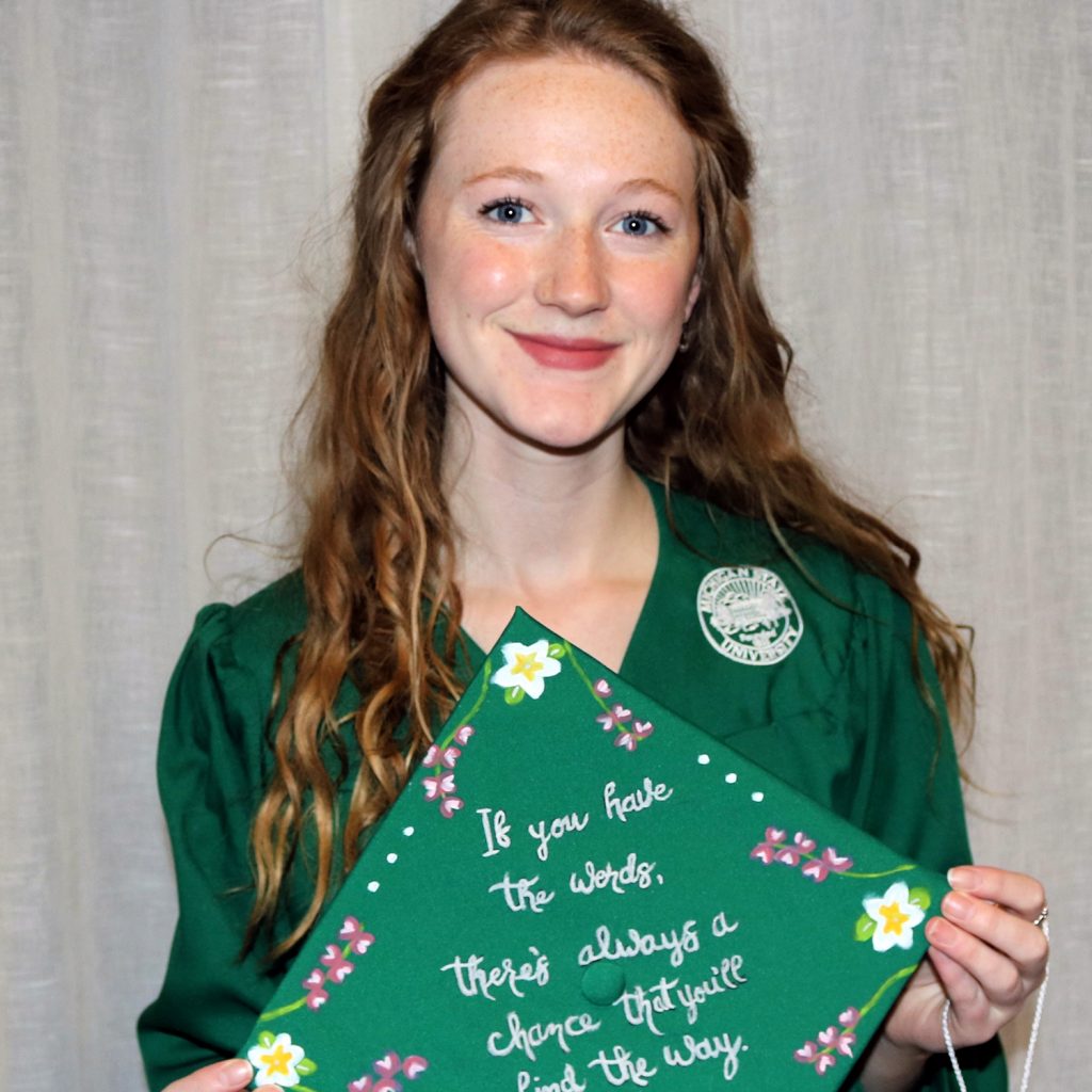 woman with red hair wearing a green graduation gown and holding a cap