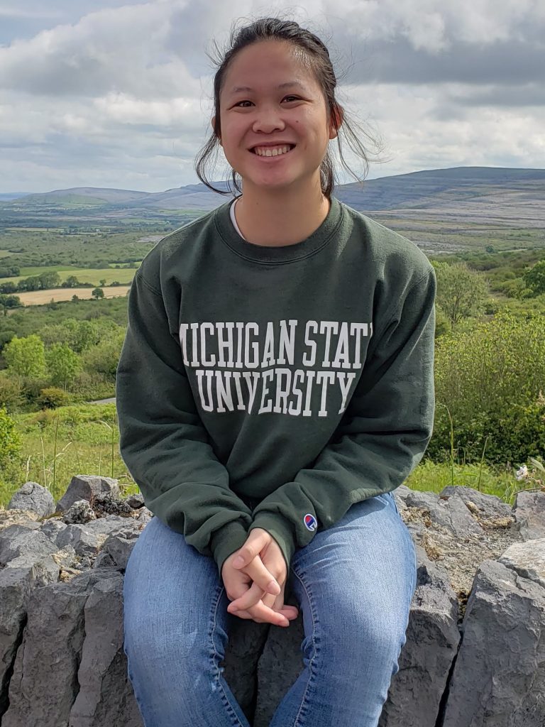 woman wearing a Michigan State University sweatshirt and jeans sitting on a stone wall