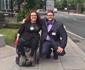 a woman with curly hair wearing a black dress and coat sitting in a wheel chair and a man in a dark grey suit with a purple shirt and tie, both wearing glasses