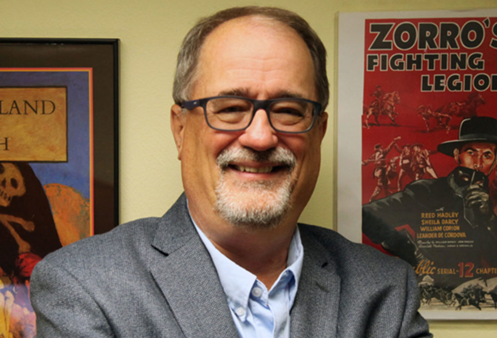 Portrait of a man in a grey suit with glasses and a goatee smiling. He poses in front of two posters.