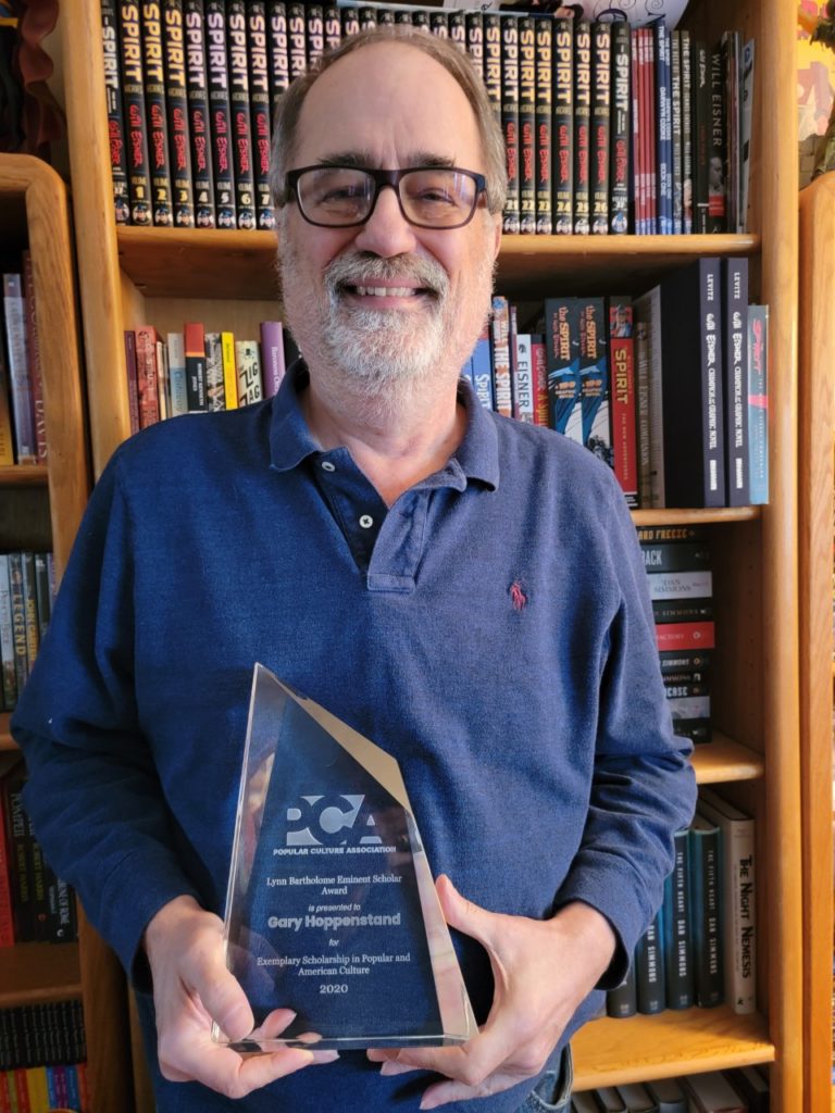 Man with black framed glasses wearing a blue dress shirt is holding an award statue that is made out of glass. He is standing in front of bookshelves.