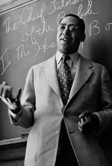 Black-and-white portrait of a man in glasses and a blazer talking in front of a chalkboard with writing on it.