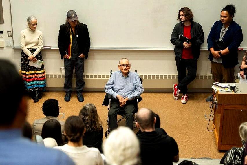 Man sitting at the front of a classroom with people gathered around listing to him talk. 