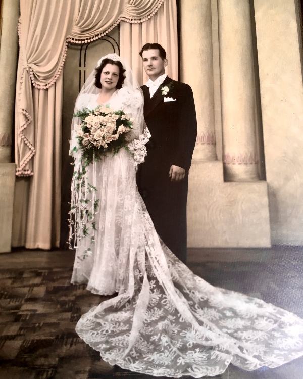 A black and white photo of a man and woman on their wedding day. The woman is wearing a wedding gown and hold a large bouquet of flowers. The man is wearing a tuxedo. 