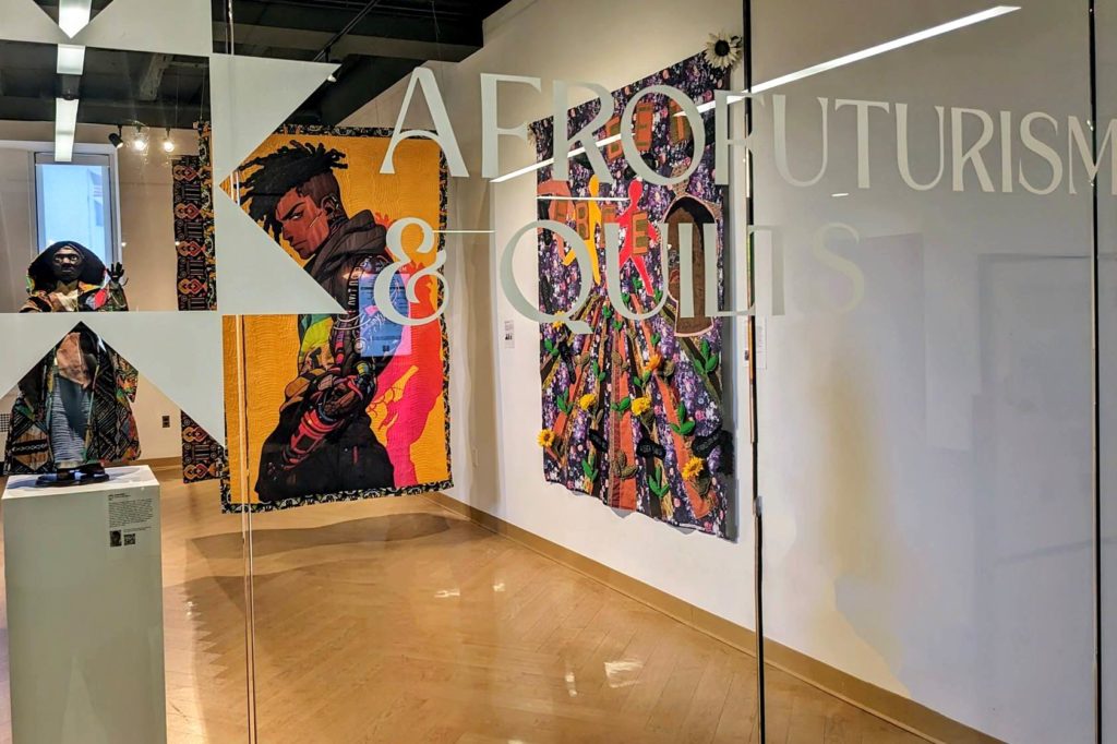 Glass wall that says: "Afrofuturism & Quilts" and you can see colorful quilts through the glass. 