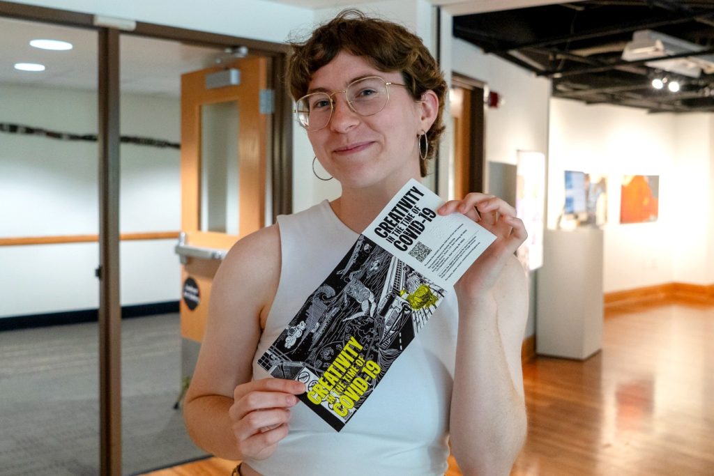 A picture of a person in a white blouse, with glasses and silver hoop earrings holding a brochure that reads "Creativity in the time of COVID"