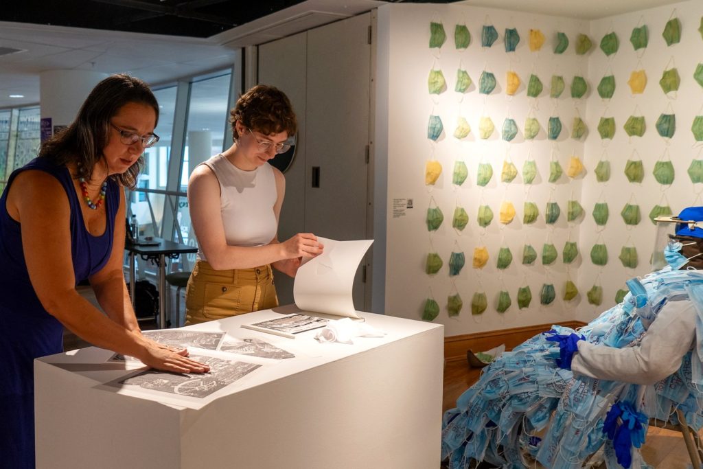 A picture of two people sorting through papers. IN the background. Someone sits on a chair, and beyond them rows of masks hang from the wall. 