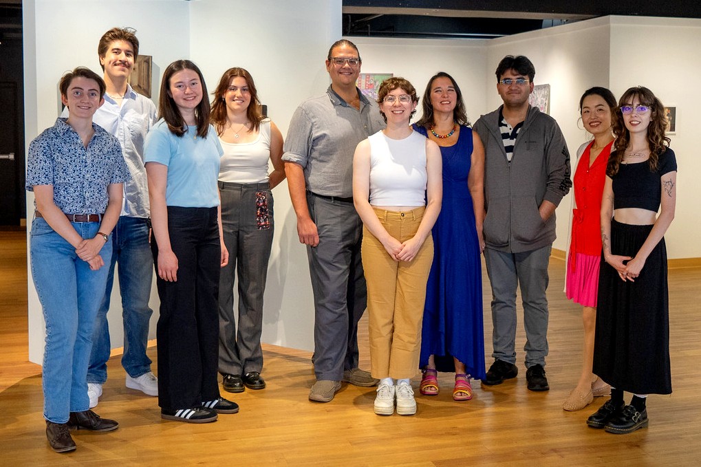 A picture of a group of people in a range of clothes standing on a wooden floor in front of white walls. 