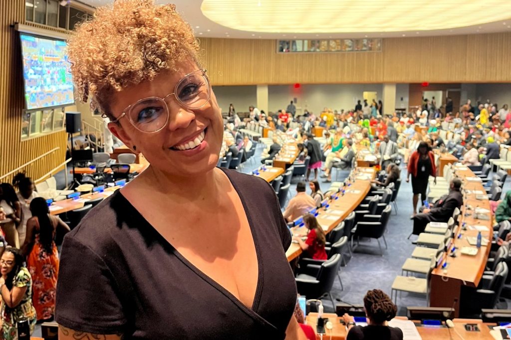 Woman with glasses and black top in the foreground with an auditorium in the background with several chairs, desks and people.