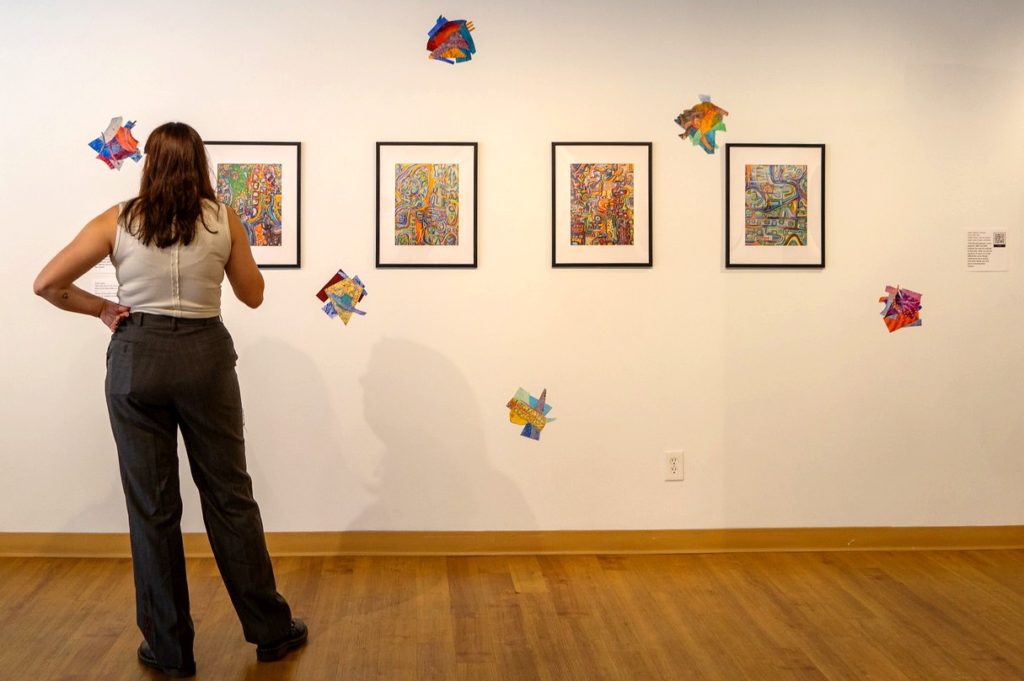 A picture of a woman standing examining four paintings. She is to the right of frame, wearing black jeans and a white top. The four paintings are multi-colored and vibrant, and colors collages are tapped to the wall around them.