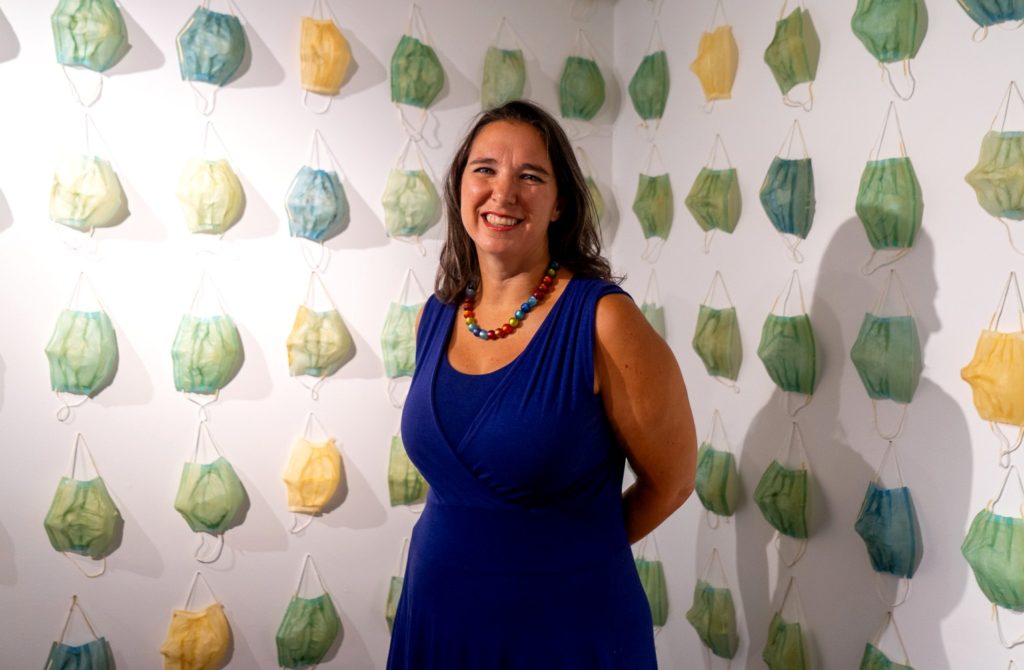 A picture of a woman in a blue dress with a band of multi-colored pearls standing in front of a wall with rows of multicolored masks hanging. 