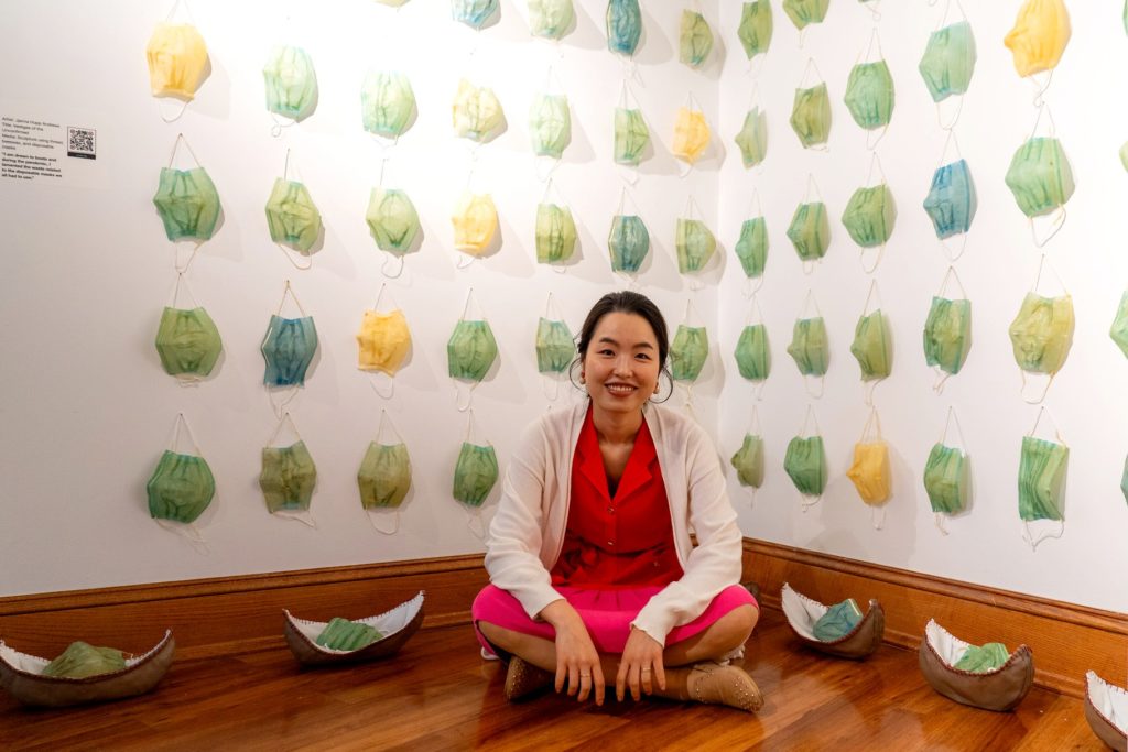 A picture of a woman in a red dress with a white cardigan sitting on the floor smiling at the camera. Behind her, rows of green, blue, and yellow masks hang on the wall.